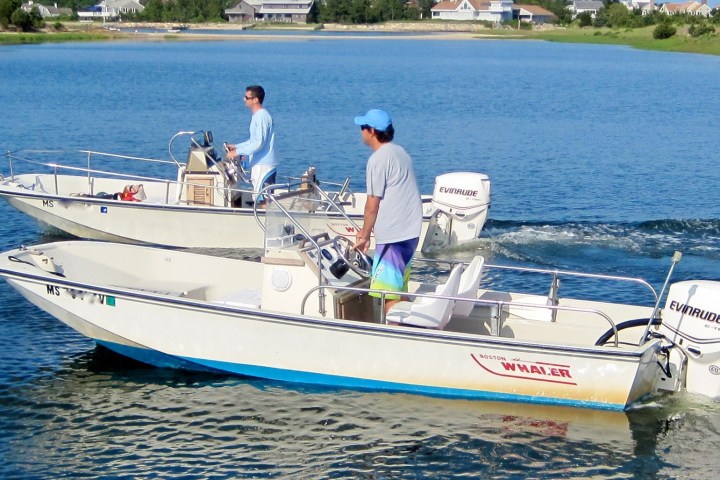 Two people operate motorboats on a calm lake near a shore with houses.