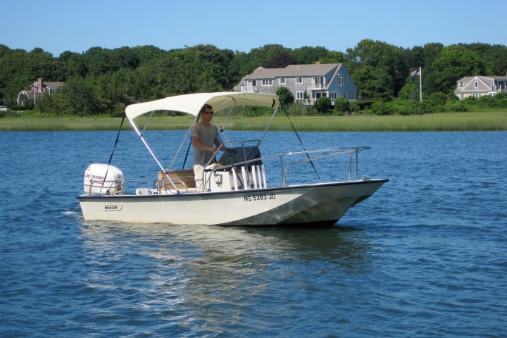 Person driving a small motorboat on a lake with houses in the background.